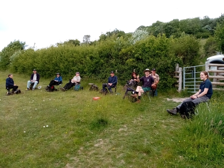 Group of volunteers sat in a semi circle on a tea break in front of a hedge and gate.