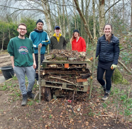 5 people stood round a bug home made of pallets and twigs