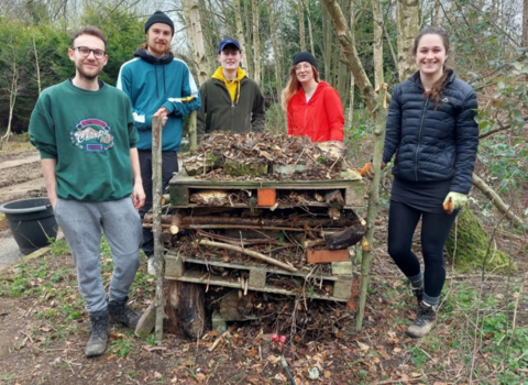 5 people stood round a bug home made of pallets and twigs