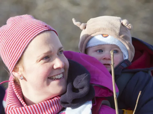 A lady in a hat and scarf carrying a baby in a hat on her back